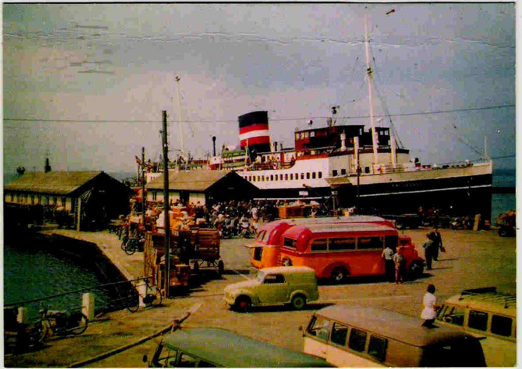 DSB-passagerskibet KALUNDBORG, bygget i 1931, ses her i Kolby Kaas havn i 1954. På kajen ses rutebiler og VW lillebiler (taxaer), der bragte passagererne til og fra skibet. Den lille rutebil er den såkaldte ”Dirch Passer-bus”. Foto: Postkort
